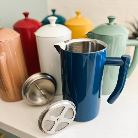 Blue ceramic pitcher with metal filter on a kitchen counter with colorful canisters in the background.