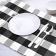 Dinner setting with a black and white checkered placemat, white plate, silver fork, knife, and spoon on a light gray surface.